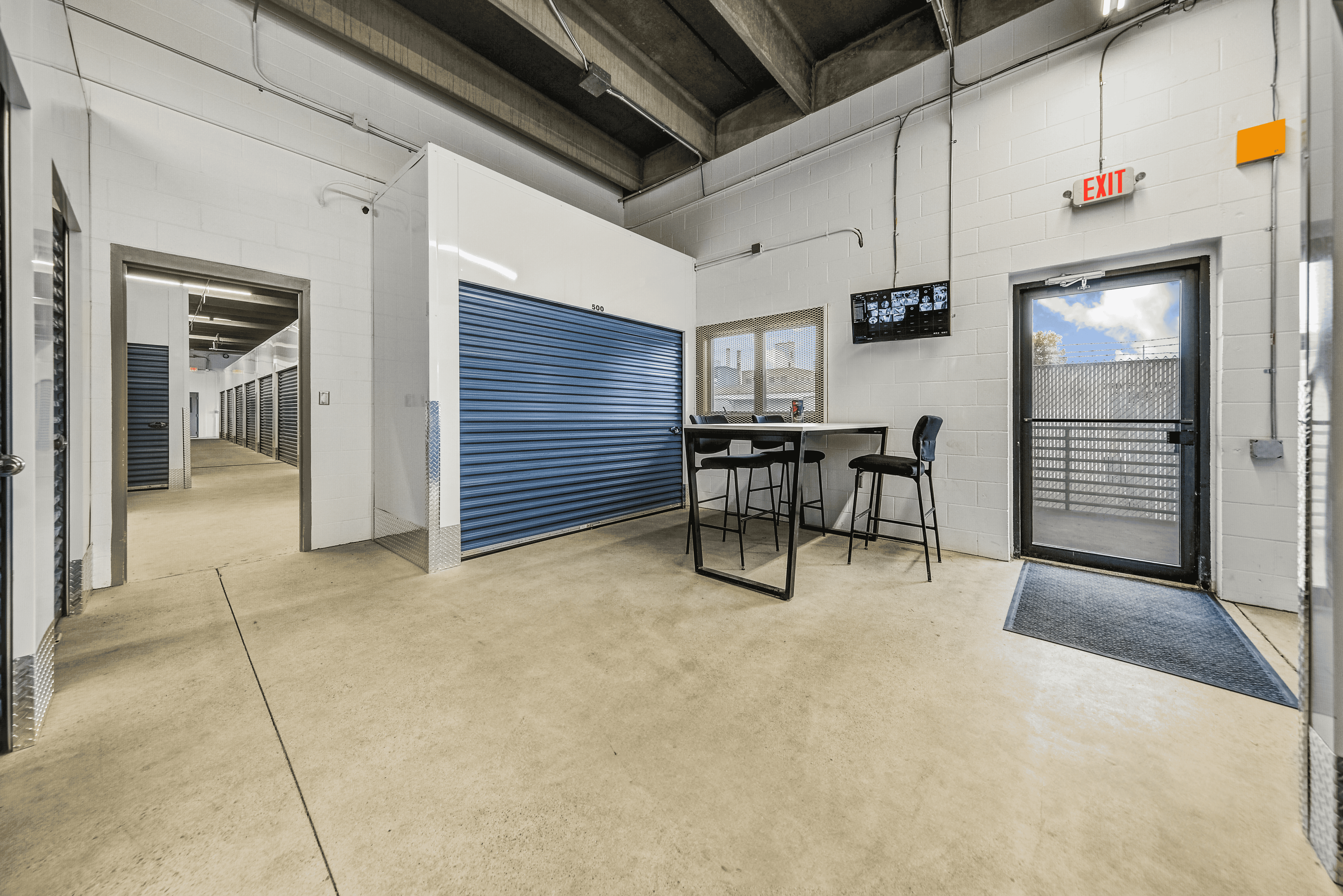 right, clean interior of Storage on 7th in Faribault, Minnesota with a wall-mounted security camera monitor and well-lit hallway of indoor storage units.
